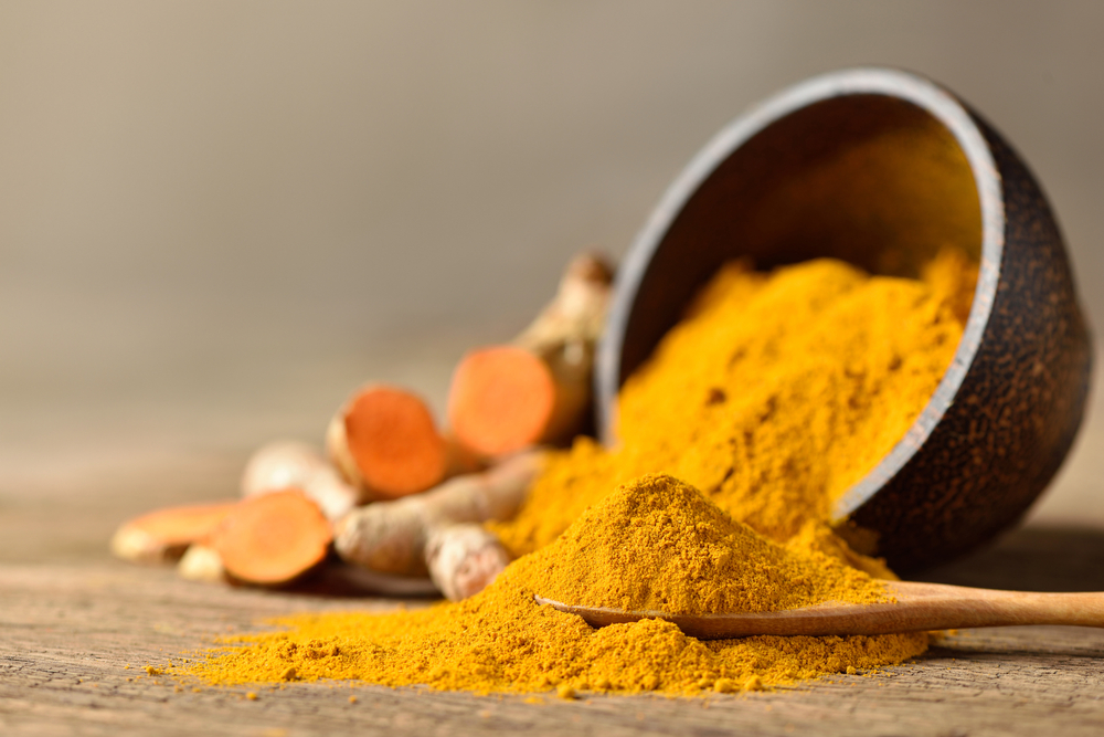 Close-up of Turmeric (curcumin) powder in wooden spoon with bowl and fresh rhizome slices on wood background.