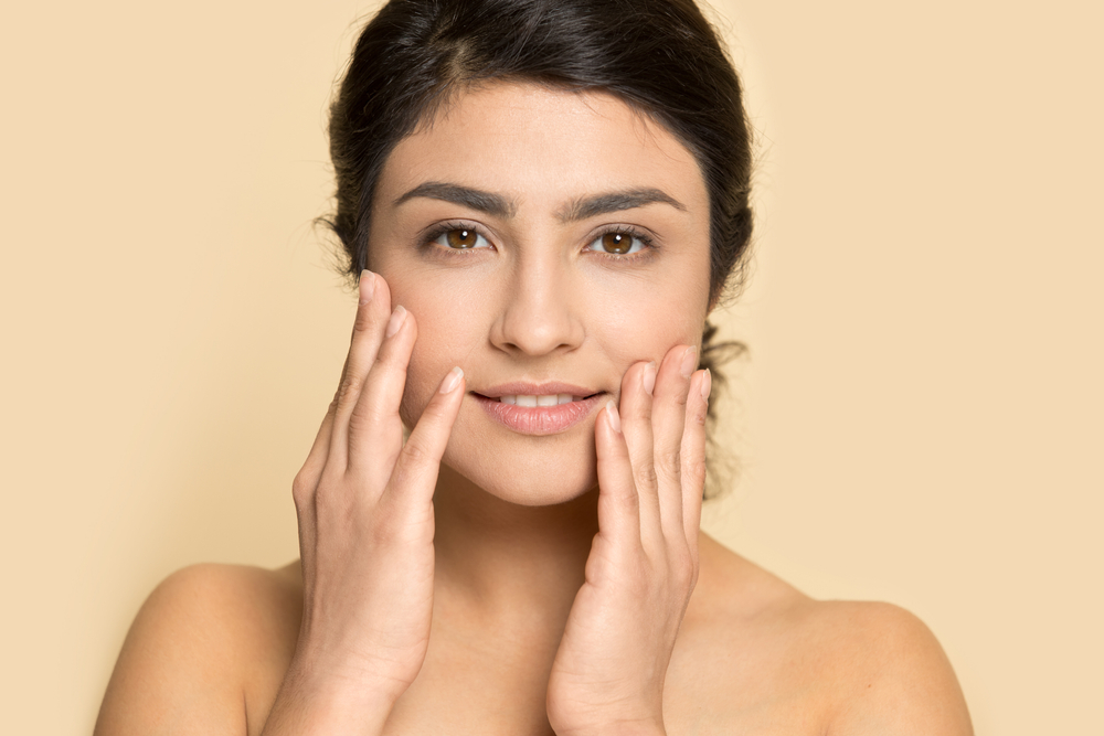 Portrait of young Indian woman isolated on yellow studio background touch healthy face skin after beauty salon procedures. Smiling multiracial female satisfied with facial treatment. Skincare concept.