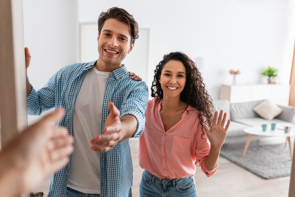 Portrait of cheerful couple inviting guests to enter home, happy young guy and lady standing in doorway of modern flat, looking out, man shaking hands, meeting new neighbors or friends
