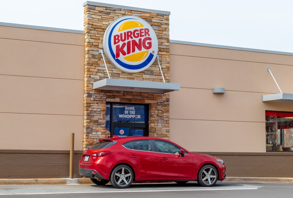 Charlotte, NCUSA - March 18, 2022: Horizontal, medium shot of "Burger King" fast food restaurant's exterior facade brand and logo sign above drive thru window with red vehicle in soft light.