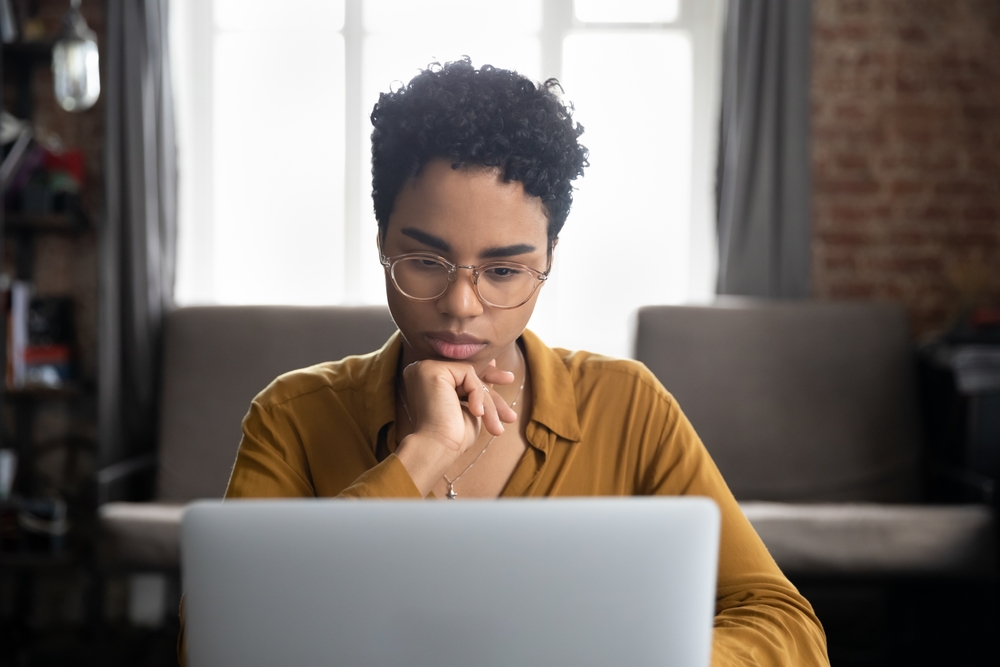 African woman in glasses sit at desk staring at laptop screen, learn new software, makes assignment, working looks concentrated, search solution or ideas. Business challenge, thinks over task concept