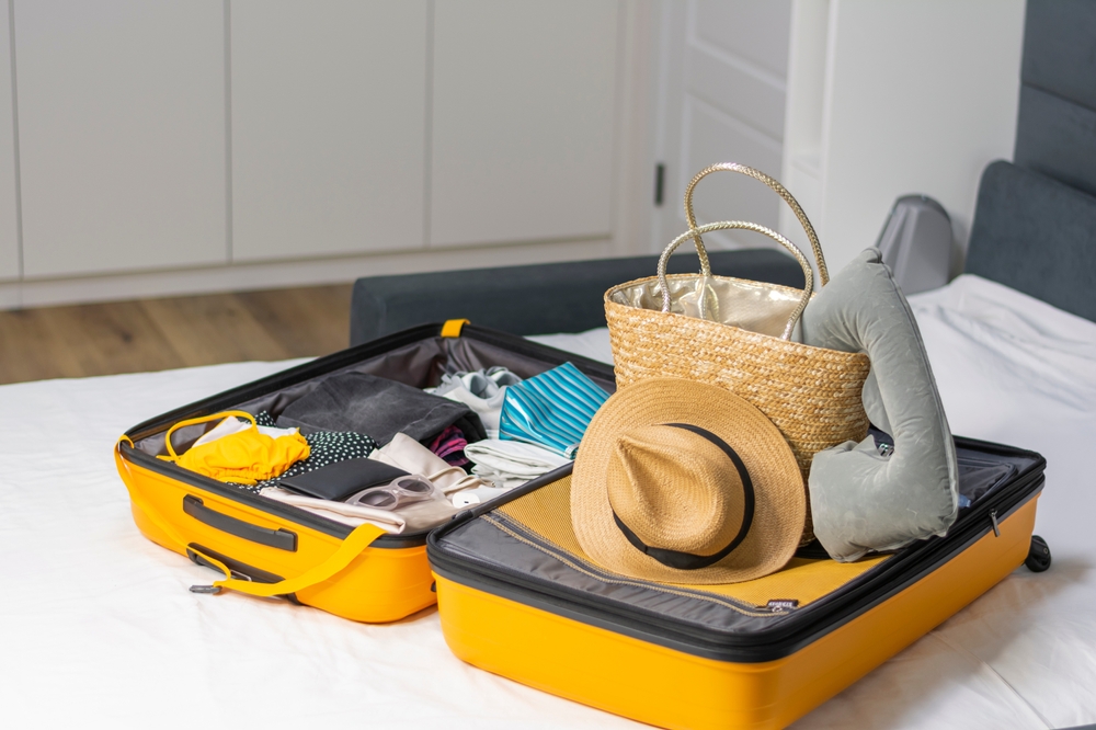 Suitcase for summer vacation on home bed or hotel room with clothes for vacation on tropical beach ocean. Straw hat and bag and swimsuit sunglasses. Concept: travel, tourism, clothing accessories