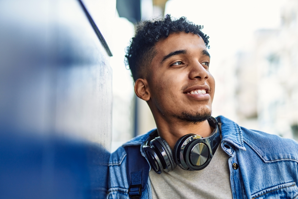 Hispanic young man smiling wearing headphones leaning on the wall