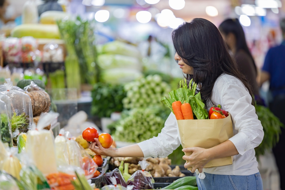 Asian woman holding large paper shopping bag of food. Woman choosing food products in grocery shop or supermarket holding vegetables in hand and thinking what to cook.