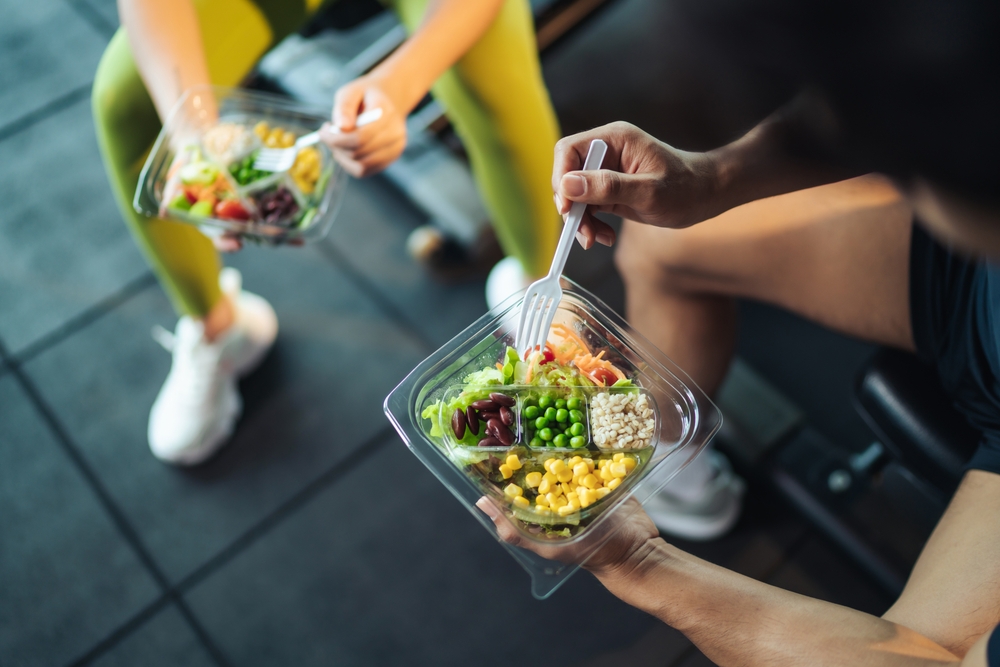 Top view Asian man and woman healthy eating salad after exercise at fitness gym. Two athlete eating salad for health together. Selective focus on salad bowl on hand.
