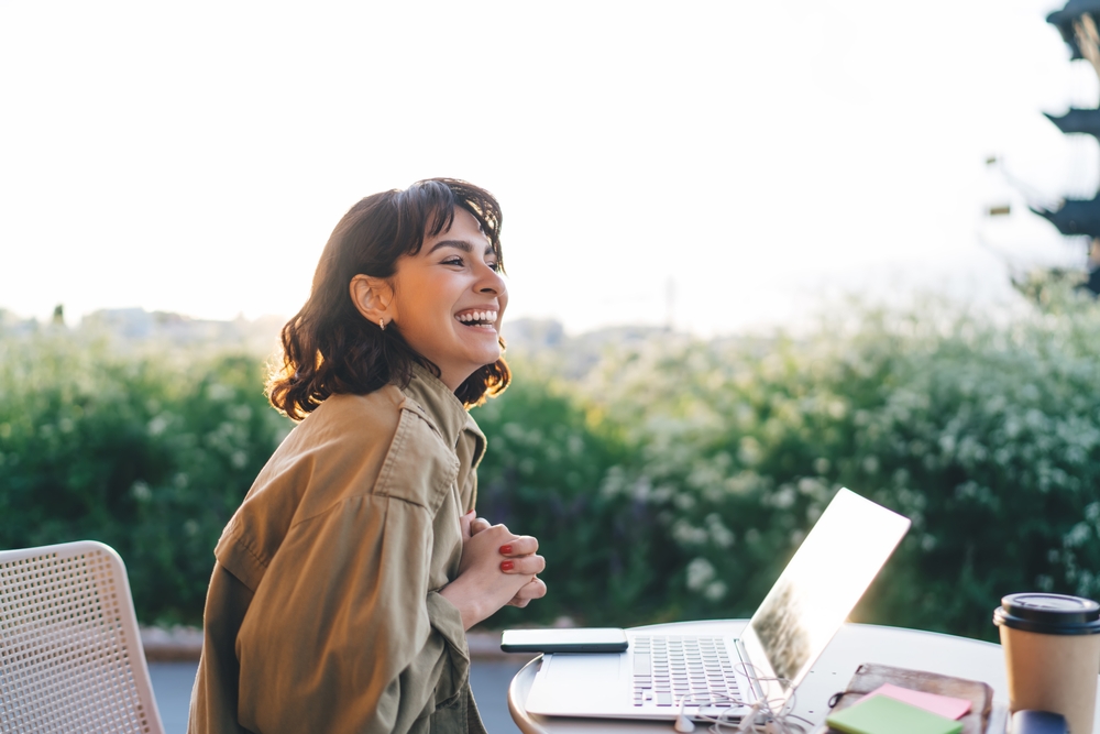 Side view of young brunette smiling happily while sitting at table with laptop working remotely on new web design project in sunny garden
