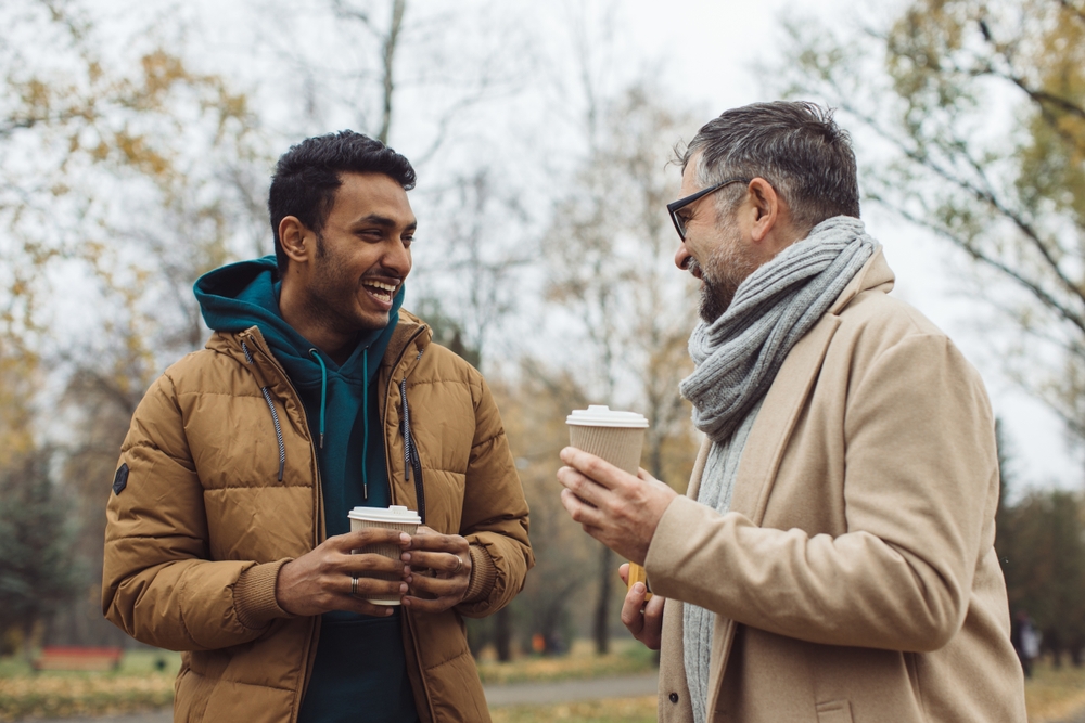 Friends, a senior and a young man walking and talking and drinking coffee together in the autumn park.