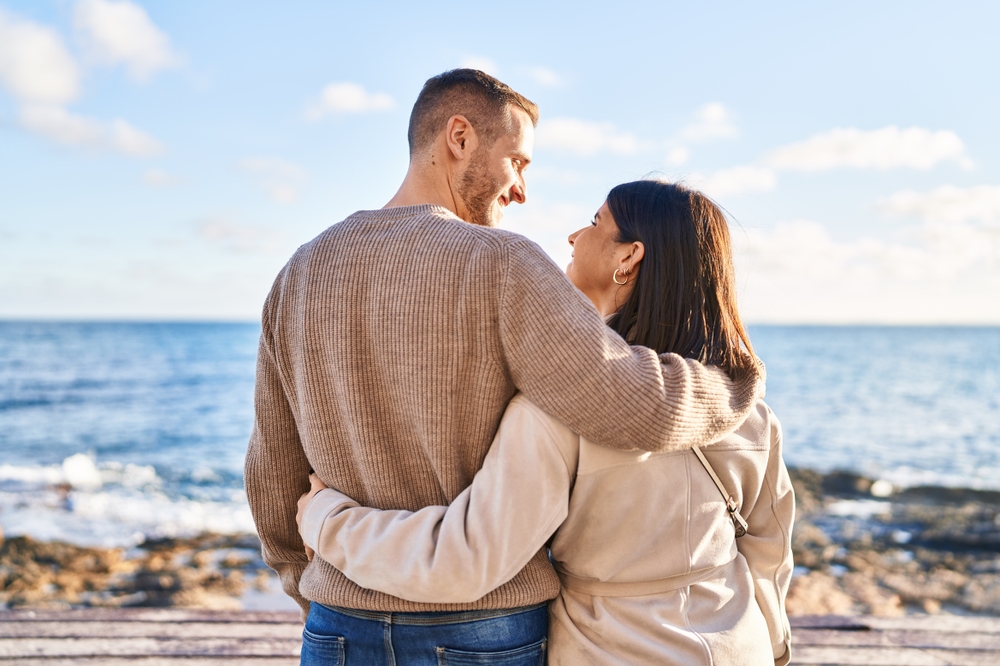 Man and woman couple hugging each other standing on back view at seaside
