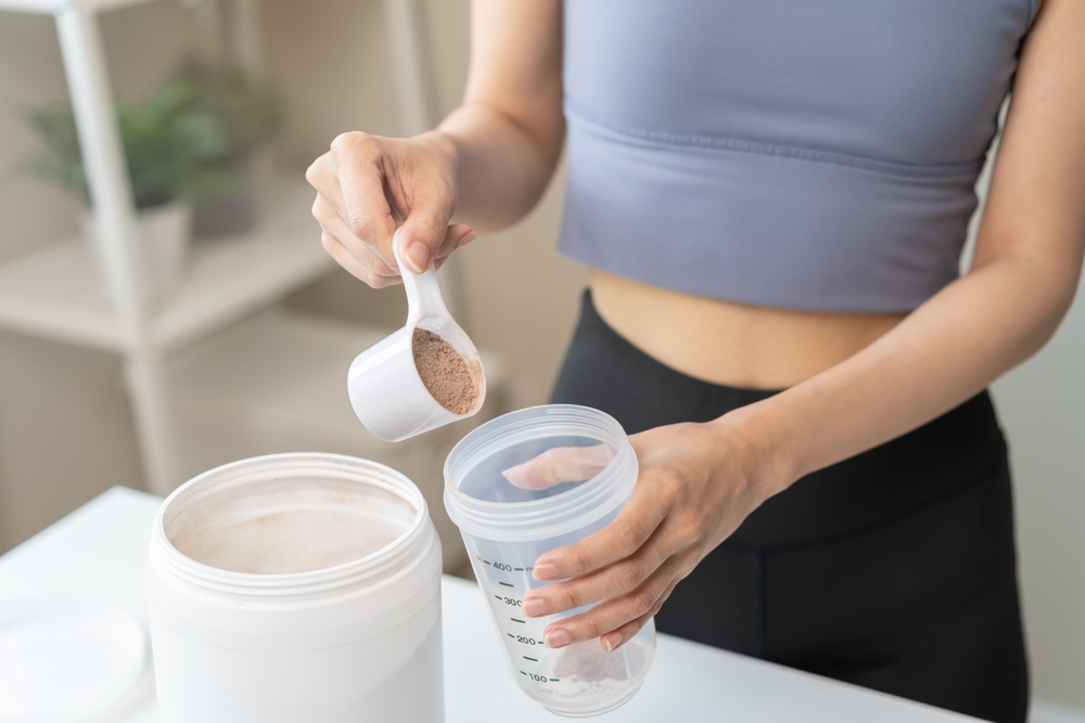 Diet meal replacement for weight loss, asian young woman in sportswear, hand in holding scoop making protein into bottle to shake, drink supplement for muscle after workout at home. Healthy body care.