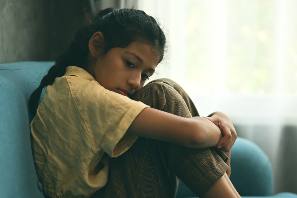 Unhappy girl portrait, Sad child sitting on sofa at home, Upset girl hugging knees alone, Concept of lonely girl and kid with trouble and violence