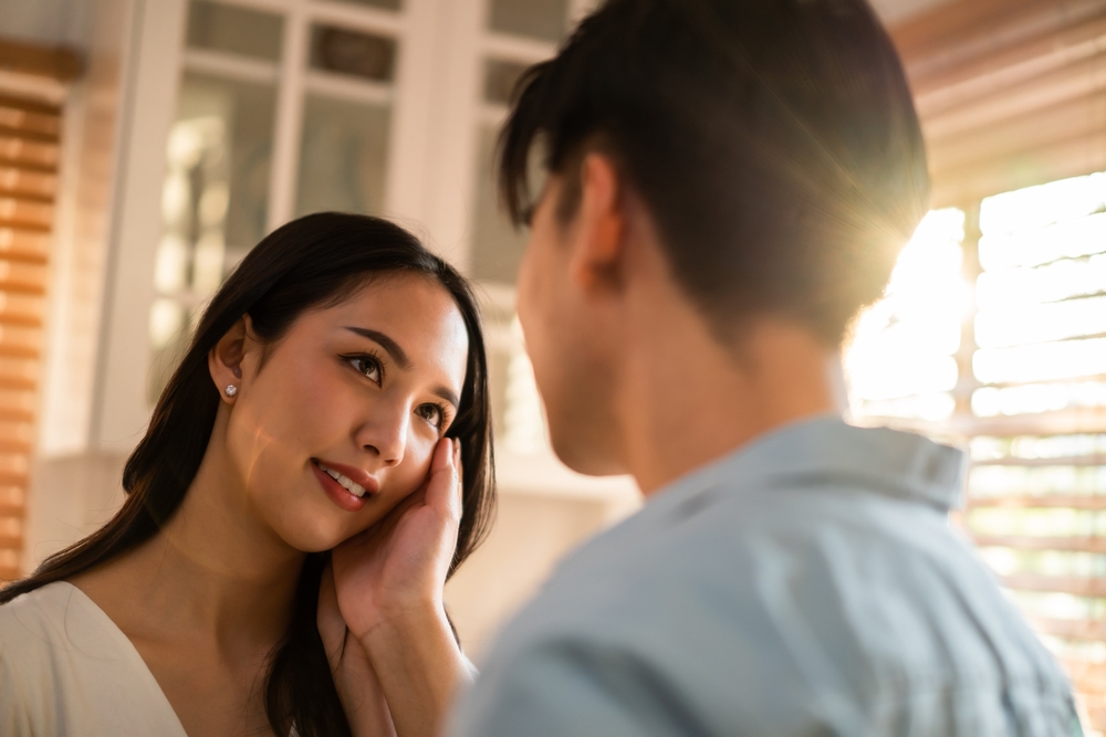 Asian young man and woman looking each other in living room at home. Attractive romantic new marriage couple male and female spend time celebrate anniversary and valentine's day together in house.
