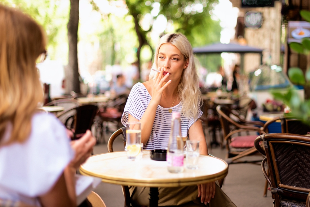 Young woman smoking a cigarette and drinking coffee while sitting outdoors at the typical french cafe terrace. Blond haired female talking with somebody.