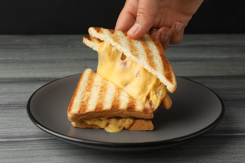 Woman taking delicious sandwich with ham and melted cheese at grey wooden table, closeup

