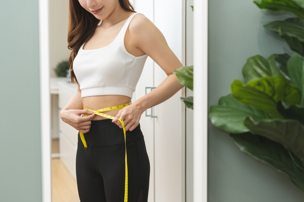 Weight lose, loss concept. Slim body asian young woman hand use tape measuring around waistline in fit sports. Girl looking reflect in mirror at home. Healthy nutrition, fitness for wellbeing beauty.