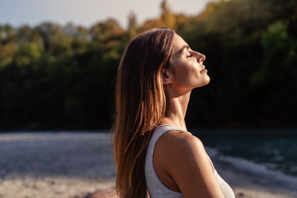 Young woman with long hair enjoying sun with closed eyes getting natural vitamin D outdoors. Peace of mind. Mindfulness, mental health, spirituality, well-being, unwind yourself
