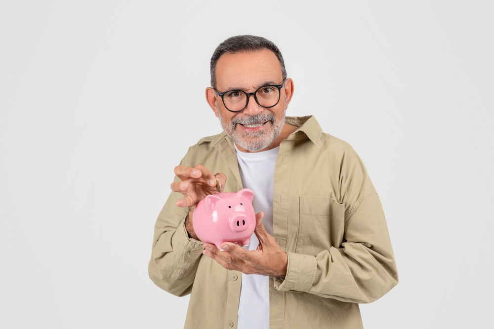 Smiling senior man holding and inserting coin into pink piggy bank, happy elderly gentleman representing smart savings and financial planning, standing on white background in studio, copy space
