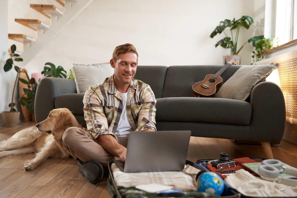 Portrait of smiling young man, tourist with a dog, packs suitcase, goes on holiday, looks at his laptop, booking a hotel or flight for the vacation.