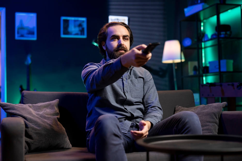 Man watching shows on TV, using remote control, paying close attention to screen. Sedentary man sitting in pajamas all day consuming mindless entertainment, picking what movie to watch next
