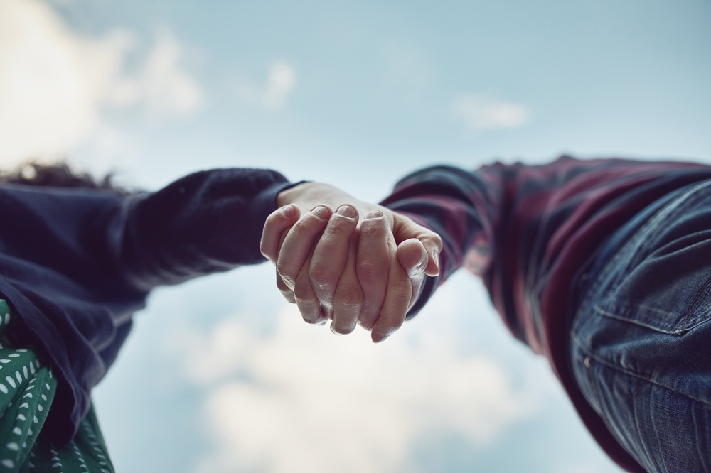 Low angle, blue sky and couple with love, holding hands and romance with adventure, relationship and bonding together. Closeup, touching palms and people with trust, marriage and nature with kindness