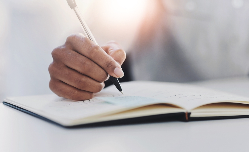 Book, hand and writing with business person closeup at desk in office for notes or planning. Agenda, appointment and schedule in diary, journal or planner at corporate workplace for profession