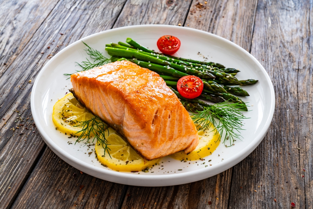 Fried salmon steak with cooked green asparagus, cherry tomatoes and lemon slices served on white plate on wooden table