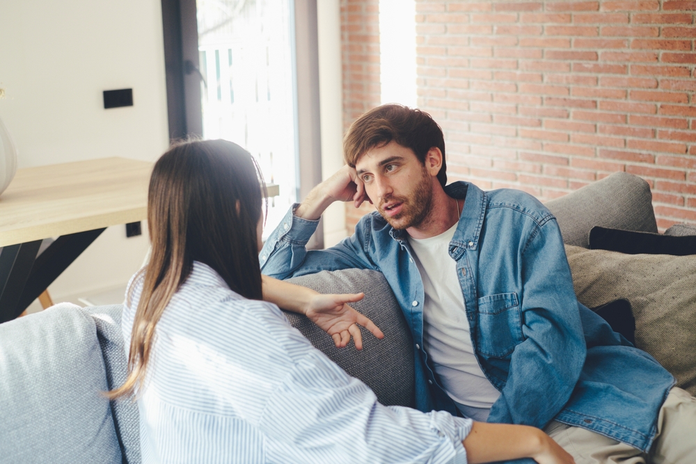Serious young couple sitting together on sofa, talking about relationships, spending time together at home, concentrated husband listening to wife talking, friends having conversation
