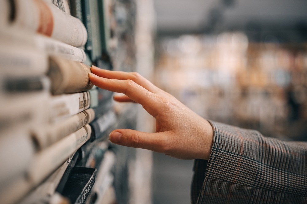 A hand reaches out to touch the spine of a book in a bookshelf, the rest of the bookstore is blurred out in the background.