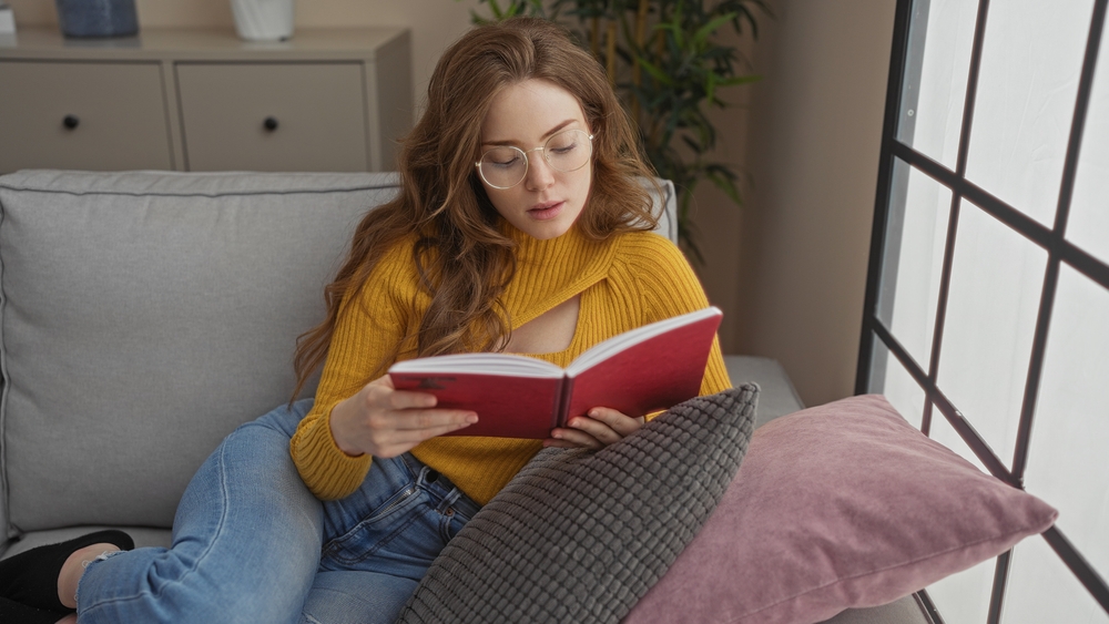 A young attractive blonde woman reading a red book in a cozy indoor living room, wearing glasses, seated on a sofa with a comfortable, relaxed atmosphere.