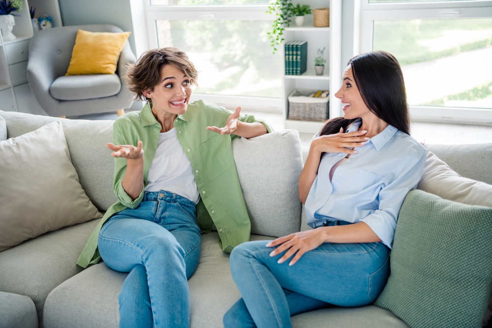 Photo of shiny impressed young girls wear shirts sitting sofa talking smiling indoors apartment room
