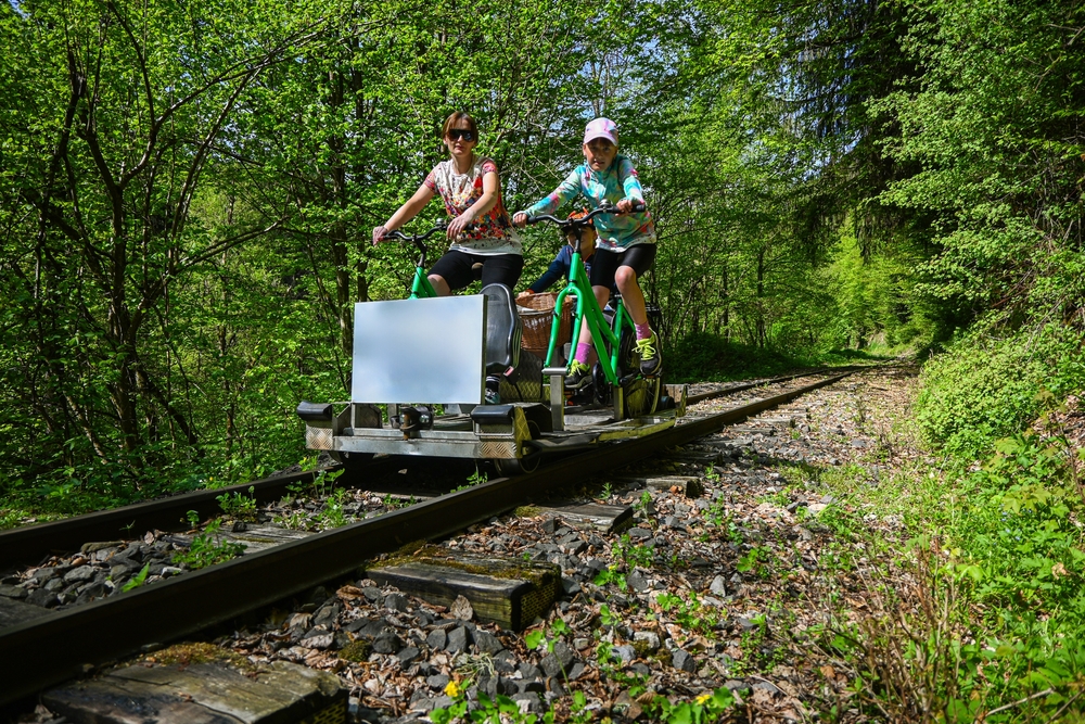 Family enjoying a sunny day, pedaling a rail-bike through a lush green forest on an old railway track.