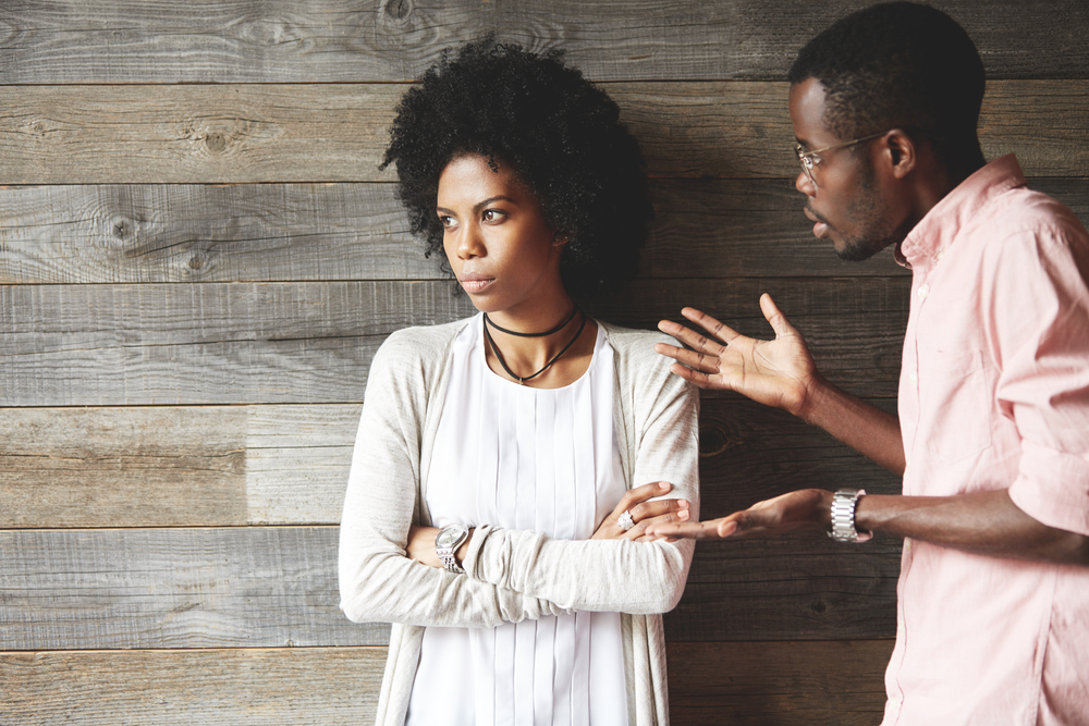 Young black indignant male gesticulating, arguing with his dark skinned girlfriend, who is standing against wooden wall with crossed arms, looking away with deeply offended expression on her face