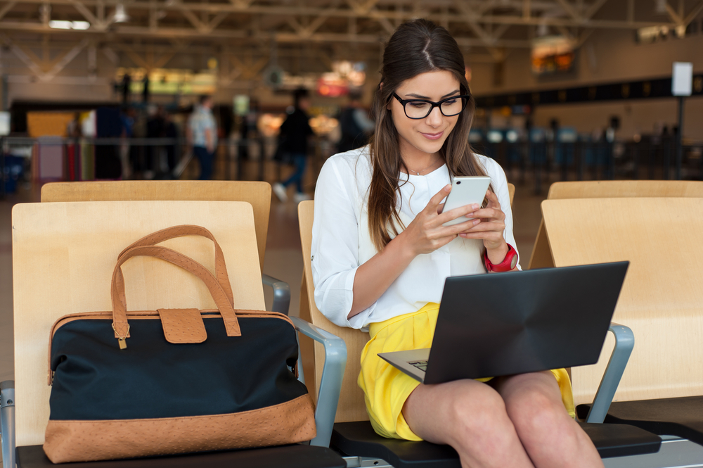 Airport Young female passenger on smart phone and laptop sitting in terminal hall while waiting for her flight. Air travel concept with young casual woman sitting with hand luggage suitcase.
