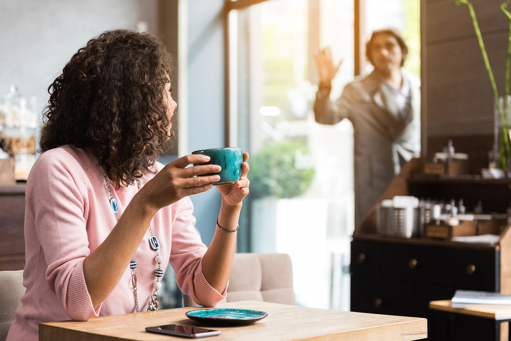 Hello. Happy young woman is meeting her friend in cafe. She is drinking coffee and sitting at table. Man standing in doors and waving hand on background