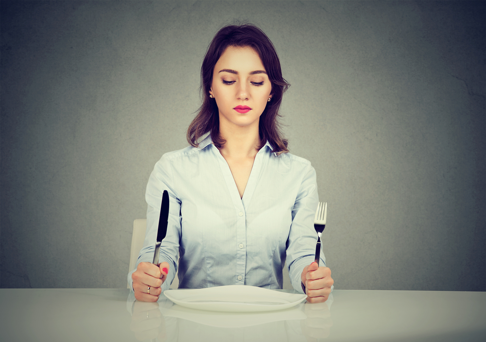 Serious woman with fork and knife sitting at table with empty plate isolated on gray wall background
