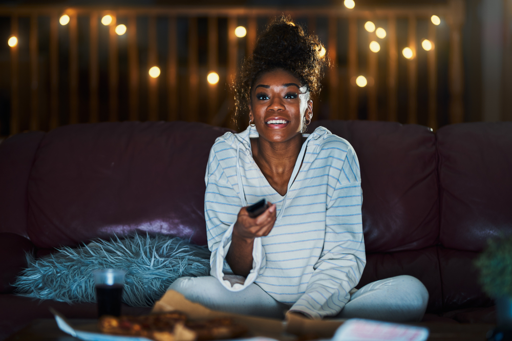 african american woman in pajamas staying up late at night eating pizza and watching tv
