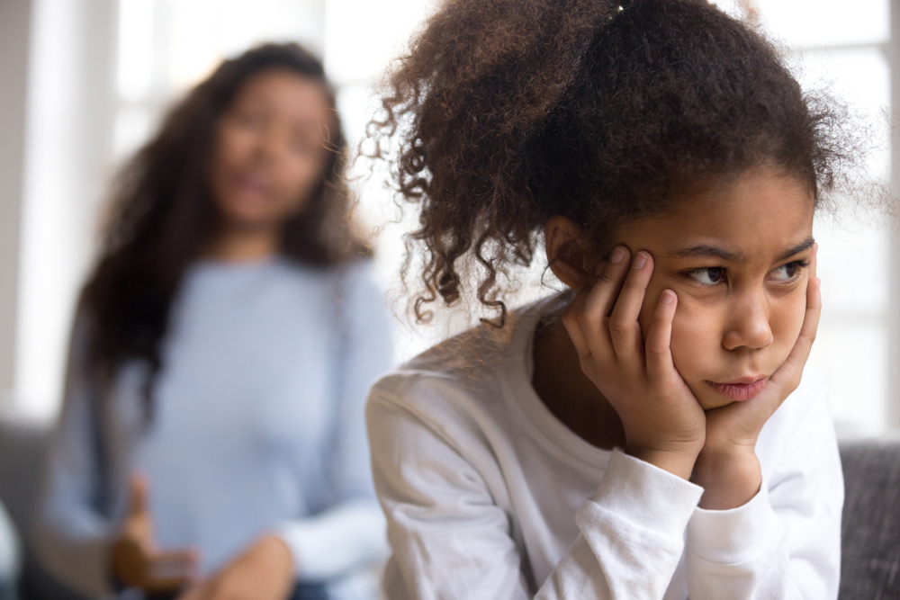 Offended African American preschooler girl looking in distance, holding head in hands, talking mother on background, family conflict, child punishment, complicated relationships with parent, close up