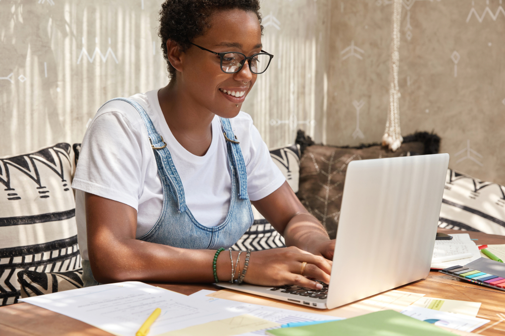 Successful young graphic designer creats something new on website, keyboards on laptop computer, chooses items in web store, makes shopping online after work with documentation. Cozy interior