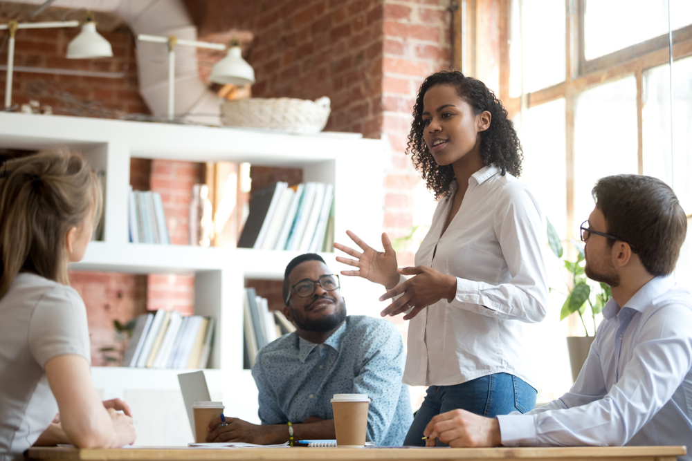 Ambitious smart african black female employee speaking at diverse meeting share creative idea opinion at group briefing while jealous envious skeptical male coworkers looking listening to colleague
