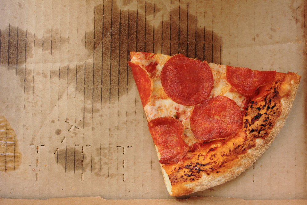 Pizza box with single slice of old pepperoni pizza in delivery box left after the party. Cold pizza leftovers in greasy cardboard, top view from above