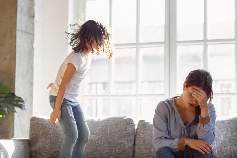 Upset mother having problem with noisy naughty daughter jumping on couch and screaming, demanding attention, frustrated mum tired of difficult child, child tantrum manipulation concept