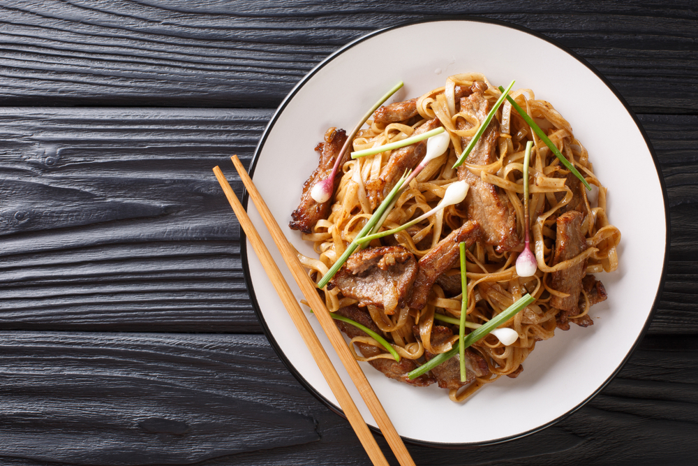 Beef Chow Fun a staple Cantonese dish, made from stir-frying beef, wide rice noodles and vegetables closeup on the plate on the wooden table. Horizontal top view from above