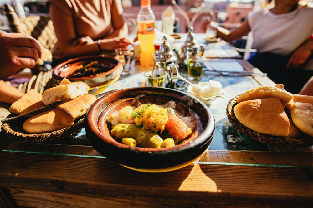 Group of people sitting down to a meal in the Medina Market in Marrakech, Marrakesch, Morocco with local foods displayed in the foreground on the table
