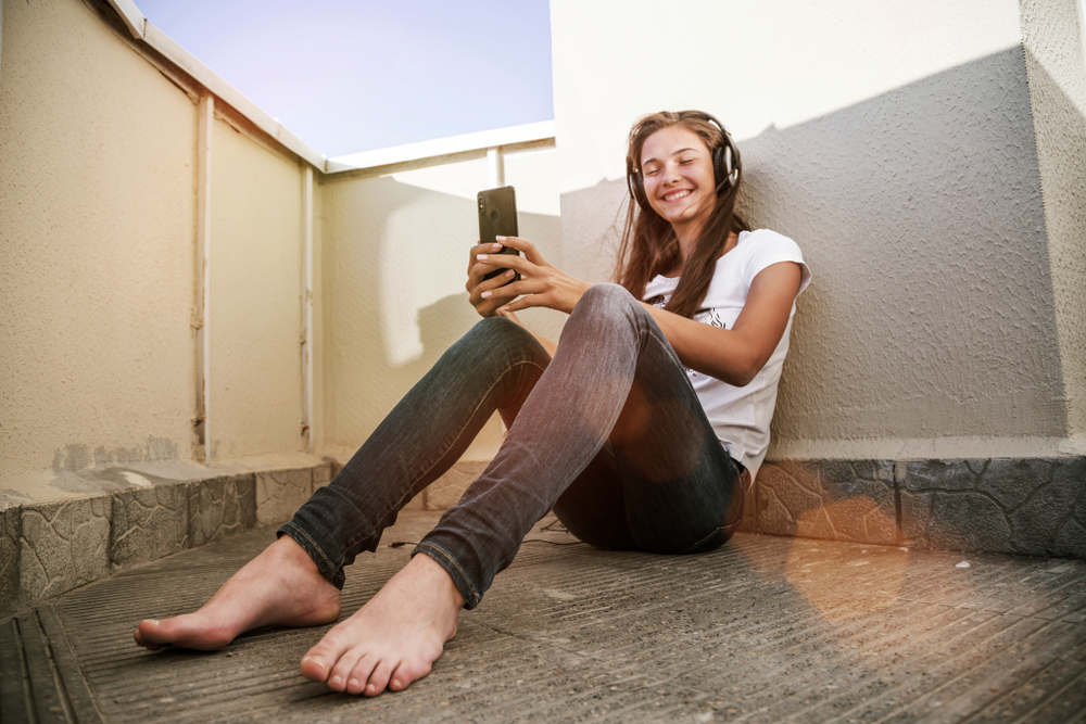 young teenager girl with smartphone and headphones sitting on floor of balcony, smilling, connecting with friends, listen to music