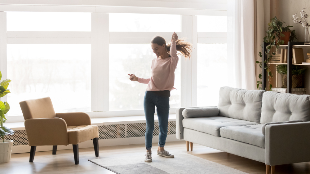 Happy carefree young woman dancing alone having fun at home listening to good music, energetic girl moving jumping in modern living room interior with large window enjoy freedom and active lifestyle