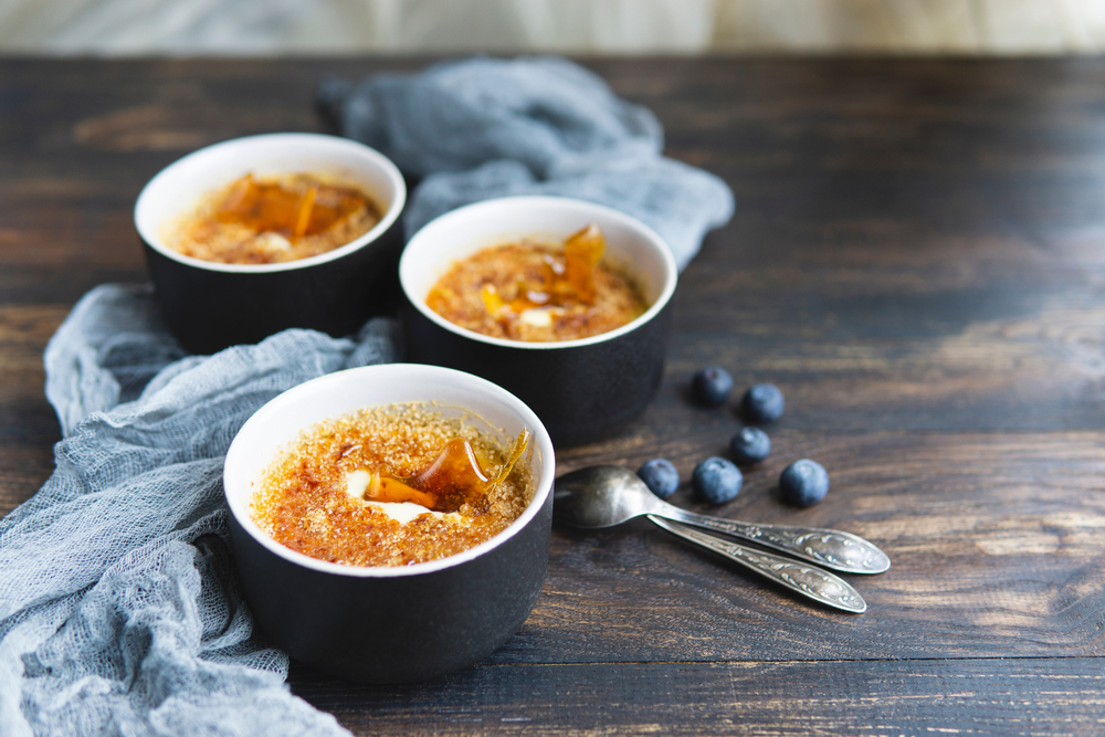 Homemade creme brulee - traditional French vanilla cream dessert with caramel decor, caramelised brown sugar on top in black baking dishes, spoons, wooden table, grey textile, blueberries. 