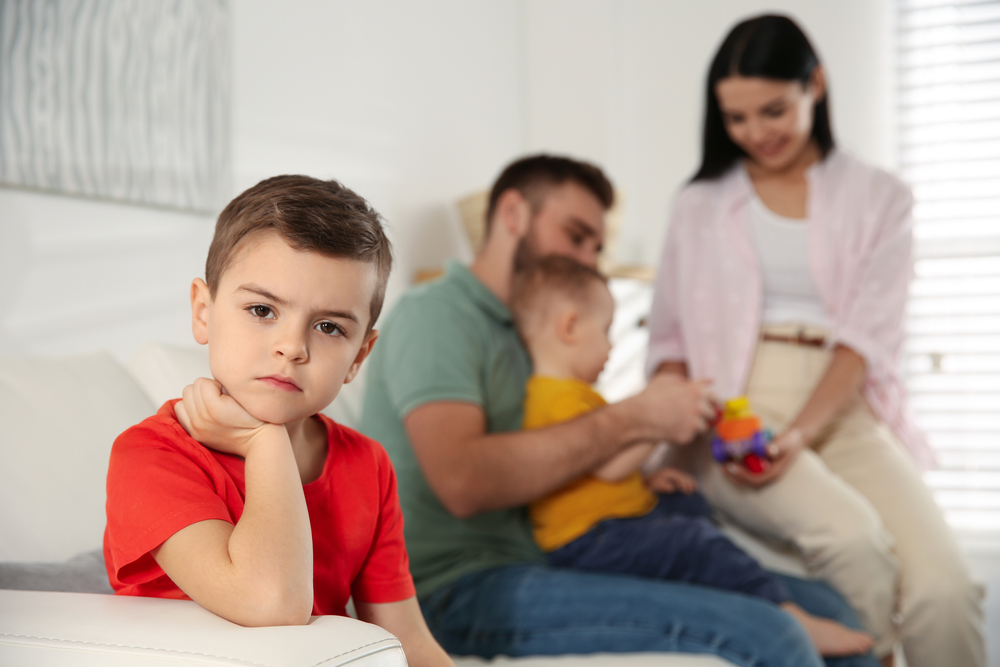Unhappy little boy feeling jealous while parents spending time with his baby brother at home