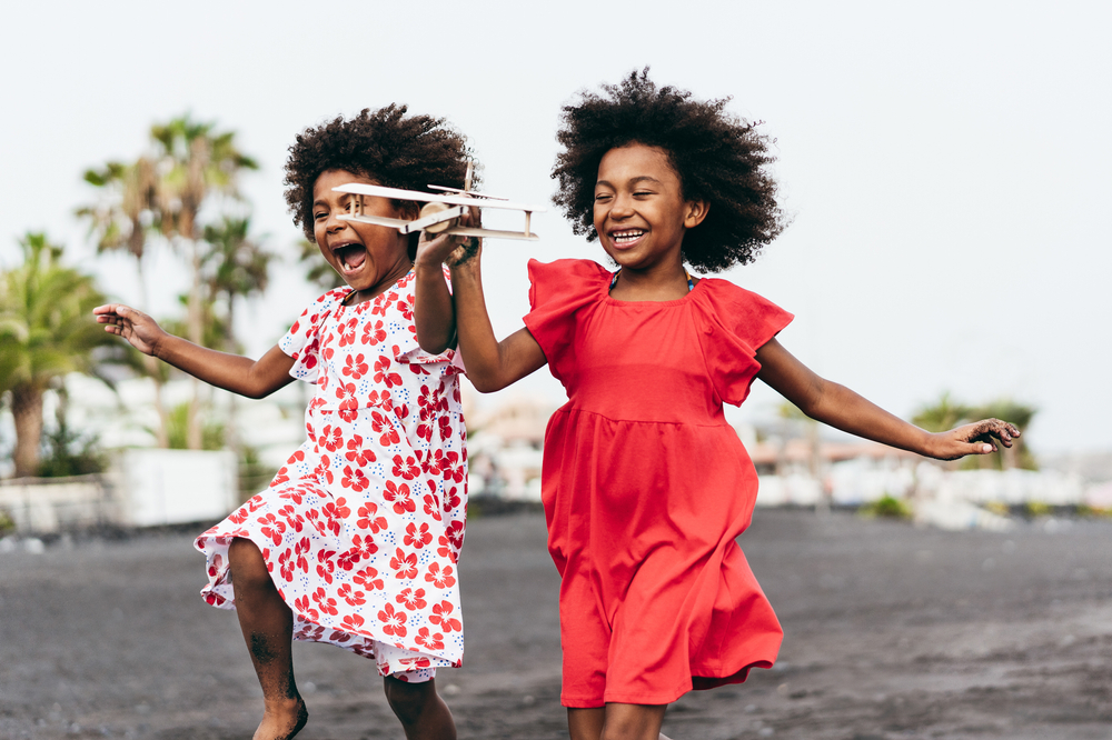Afro twins sisters running on the beach while playing with wood toy airplane - Youth lifestyle and travel concept - Main focus on right kid face
