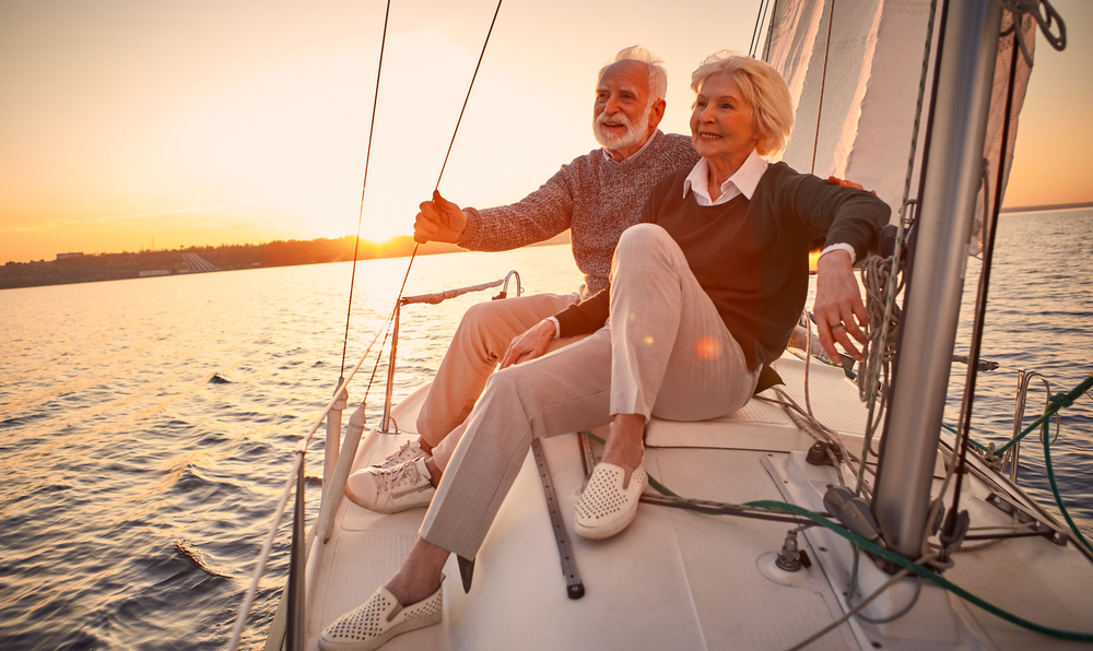 Beautiful and happy senior couple in love sitting on the side of sailboat or yacht deck floating in sea at sunset and enjoying amazing view, sailing together