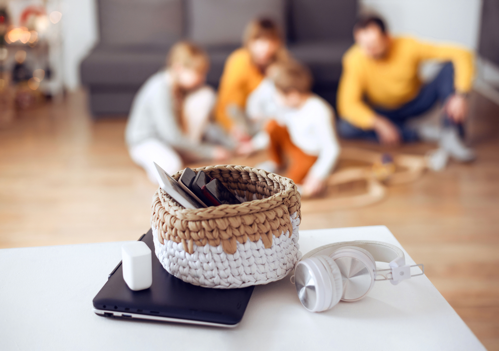 Mobile phones, laptops and digital tablets in a basket on table at home with playing family at back. Spend weekend together without technology.