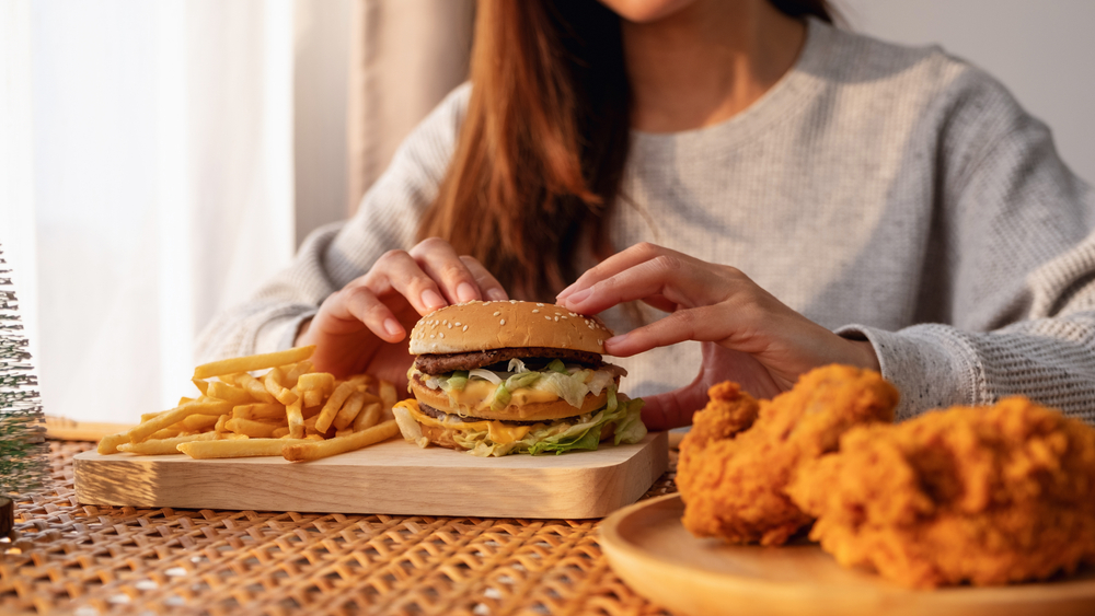Closeup image of a woman holding and eating hamburger and french fries with fried chicken on the table at home